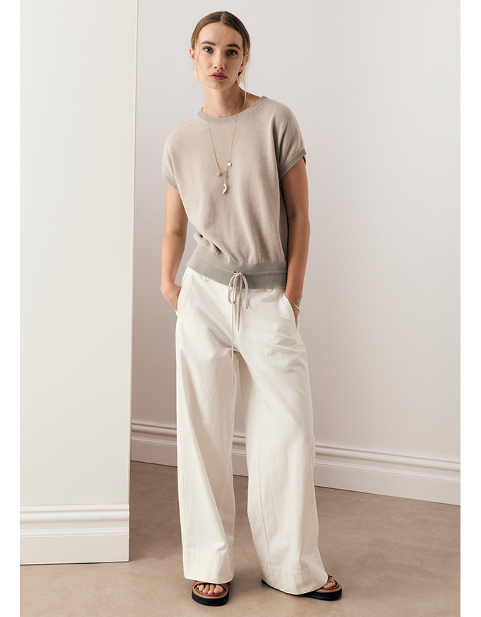 A woman modeling a beige short-sleeve knit top with a gray waistband detail, paired with off-white wide-leg drawstring pants and black sandals, standing in a minimalist room with white walls and a beige floor.