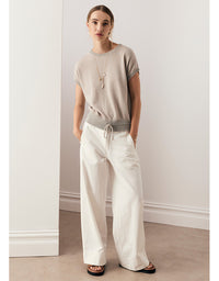 A woman modeling a beige short-sleeve knit top with a gray waistband detail, paired with off-white wide-leg drawstring pants and black sandals, standing in a minimalist room with white walls and a beige floor.