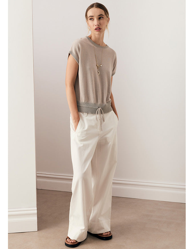 A woman modeling a taupe short-sleeve knit top with a gray drawstring waist and off-white wide-leg pants, standing in a minimalist room with beige walls and a light wood floor.
