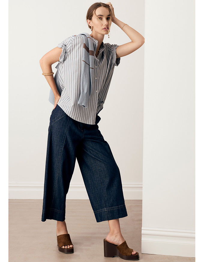 A woman models a short-sleeve button-up shirt with vertical blue and white stripes, styled with dark wide-leg cropped pants and brown platform mule sandals in a minimalist indoor setting.