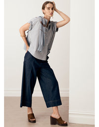 A woman models a short-sleeve button-up shirt with vertical blue and white stripes, styled with dark wide-leg cropped pants and brown platform mule sandals in a minimalist indoor setting.