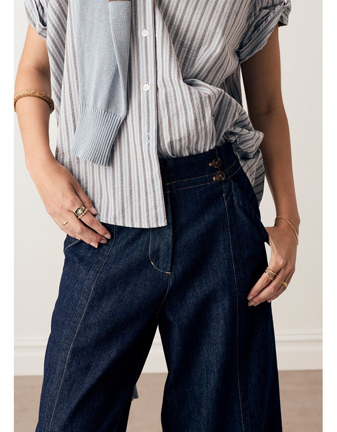 A person wearing dark blue wide-leg denim jeans with a striped button-up shirt and a light blue knit sweater draped off the shoulder, standing indoors against a light neutral wall.