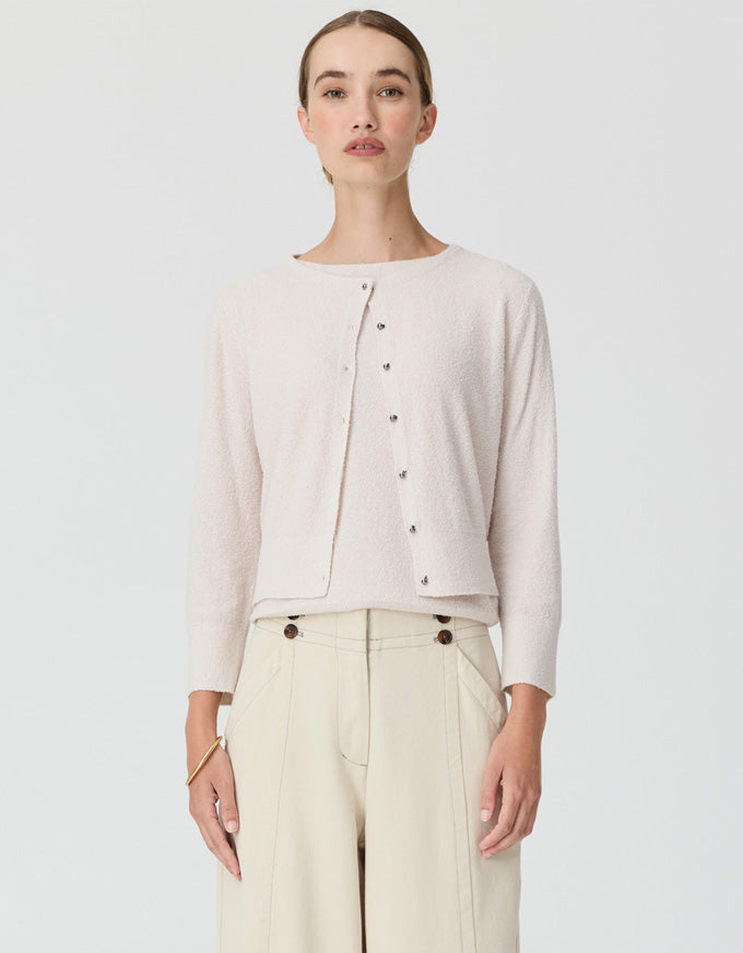 A woman modeling a light beige knit cardigan with a buttoned front and a relaxed fit, paired with cream wide-leg trousers, against a solid light gray background.
