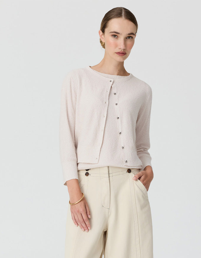 A woman modeling a light beige cropped cardigan with a button-up front, paired with cream-colored trousers, against a solid light gray background.