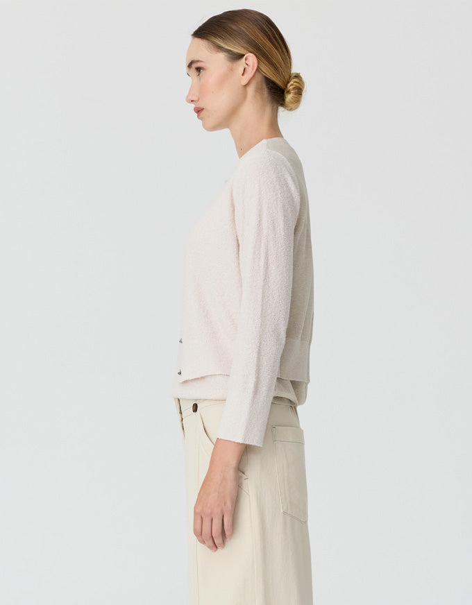 A woman shown in side profile wearing a light cream knit cardigan and matching cream trousers against a plain light gray studio background.