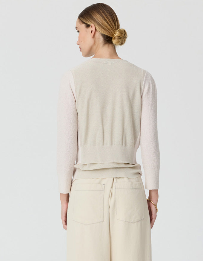 A woman shown from the back wearing a light beige knit cardigan over a pale pink long-sleeve top and cream wide-leg trousers, against a light gray studio background.