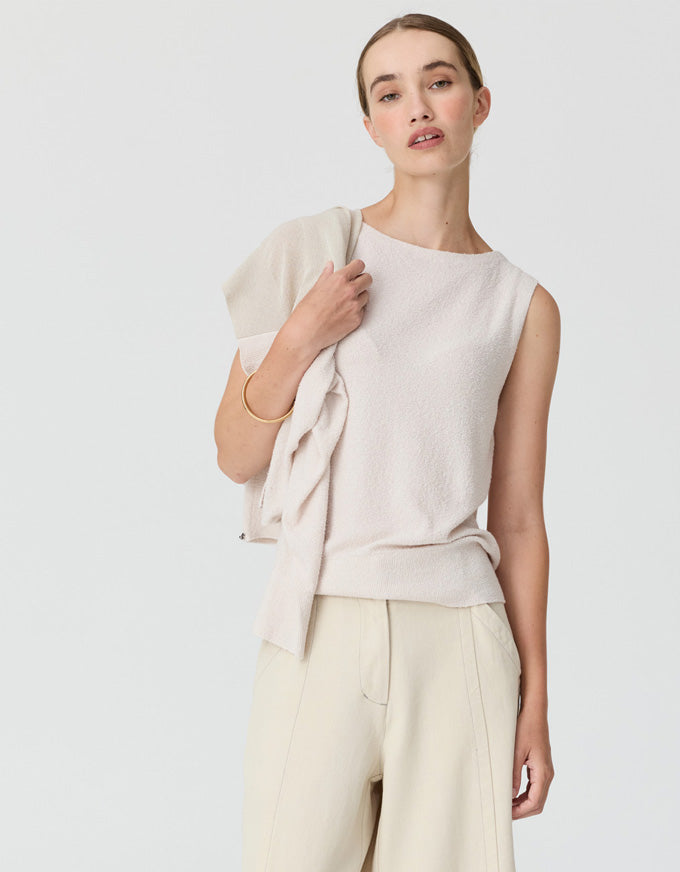 A woman modeling a sleeveless knit top in a light cream color, styled with matching wide-leg trousers and a draped cardigan, against a plain light gray studio background.