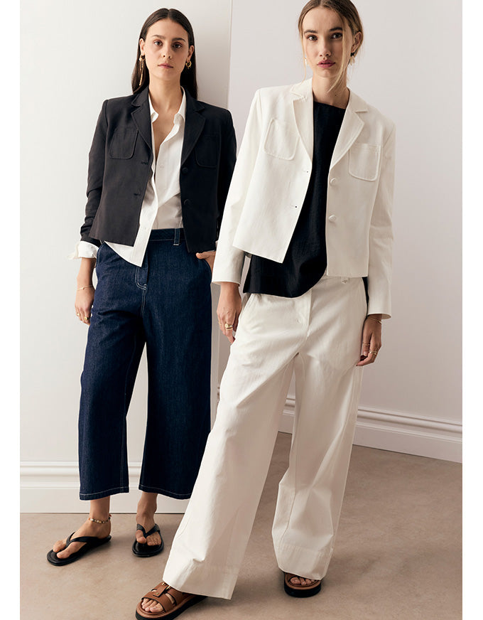 Two women modeling tailored jackets and wide-leg trousers in a minimalist indoor setting. One wears a black cropped jacket with white shirt and dark blue cropped pants, and the other wears a white cropped jacket with a black top and off-white wide-leg pants.