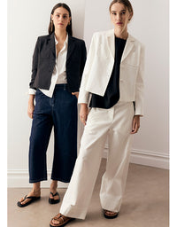 Two women modeling tailored jackets and wide-leg trousers in a minimalist indoor setting. One wears a black cropped jacket with white shirt and dark blue cropped pants, and the other wears a white cropped jacket with a black top and off-white wide-leg pants.