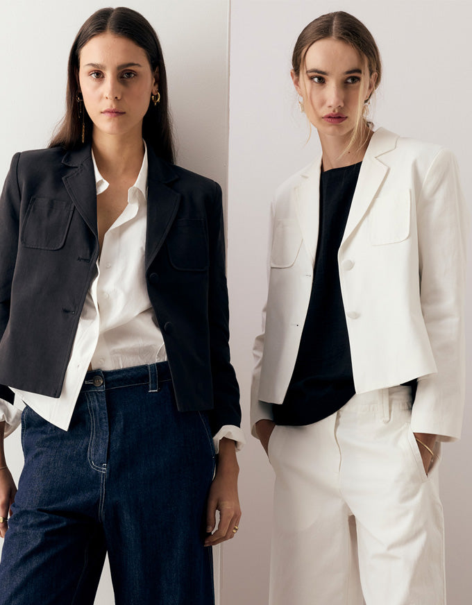 Two models posing in tailored blazer outfits against a clean off-white background. The model on the left wears a dark charcoal blazer over a white shirt with dark blue jeans.
