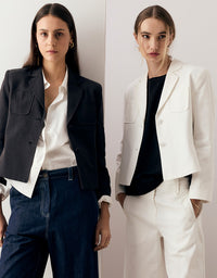 Two models posing in tailored blazer outfits against a clean off-white background. The model on the left wears a dark charcoal blazer over a white shirt with dark blue jeans.