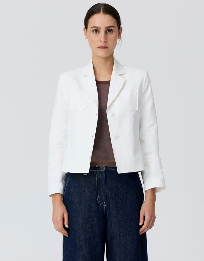 A woman modeling a white cropped blazer with notch lapels and flap chest pockets, worn over a brown top and dark blue wide-leg jeans, against a light gray studio background.