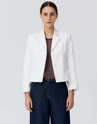 A woman modeling a white cropped blazer with notch lapels and flap chest pockets, worn over a brown top and dark blue wide-leg jeans, against a light gray studio background.
