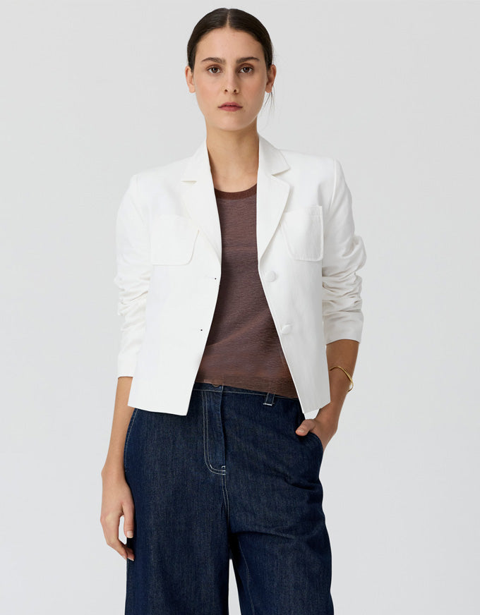 A woman modeling a white cropped blazer over a brown top and dark blue jeans, standing against a light gray studio background.