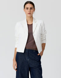 A woman modeling a white cropped blazer over a brown top and dark blue jeans, standing against a light gray studio background.
