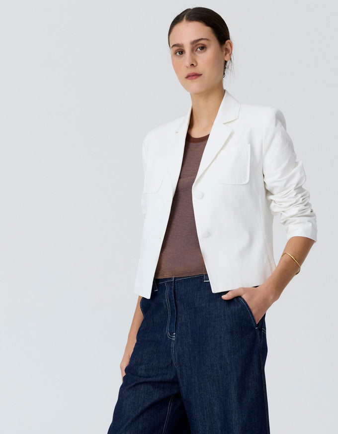 A woman modeling a white cropped blazer over a brown top and dark blue jeans, standing against a light gray studio background.