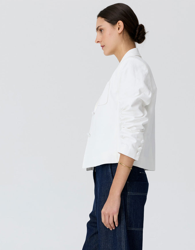 A woman shown in profile wearing a white cropped blazer with three-quarter sleeves and dark blue jeans, photographed against a clean light gray studio background.