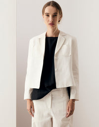 A woman modeling a cropped white blazer with notch lapels and flap chest pockets over a black top, paired with white trousers, against a minimalist off-white background.