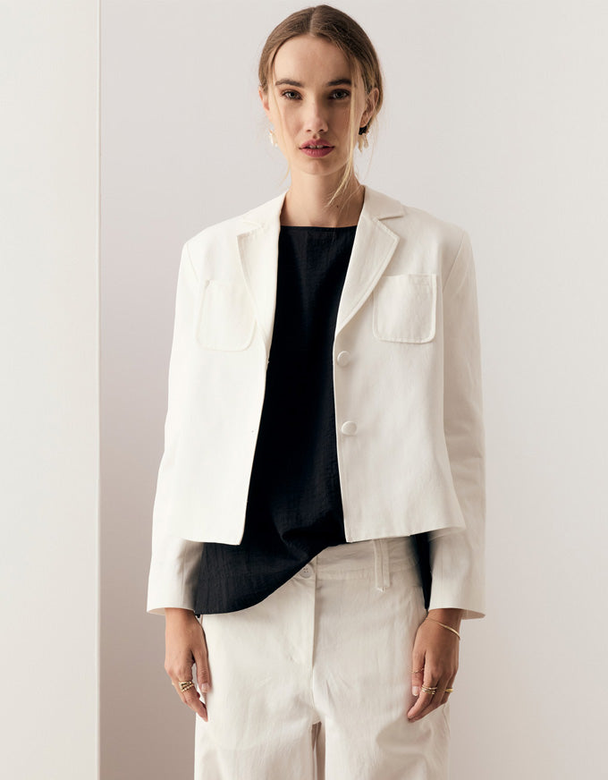 A woman modeling a cropped white blazer with notch lapels and flap chest pockets over a black top, paired with white trousers, against a minimalist off-white background.