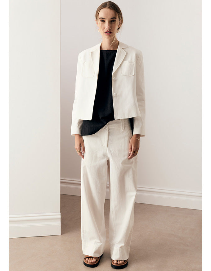 A woman stands indoors wearing a white cropped blazer, white wide-leg pants, a black top, and black sandals. The photo is taken in a minimal room with light walls and a beige floor.