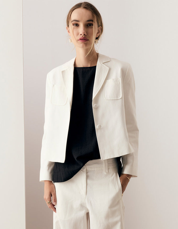 A woman modeling a white cropped blazer with notch lapels and flap chest pockets, worn over a black top and paired with white trousers, against a plain light background.