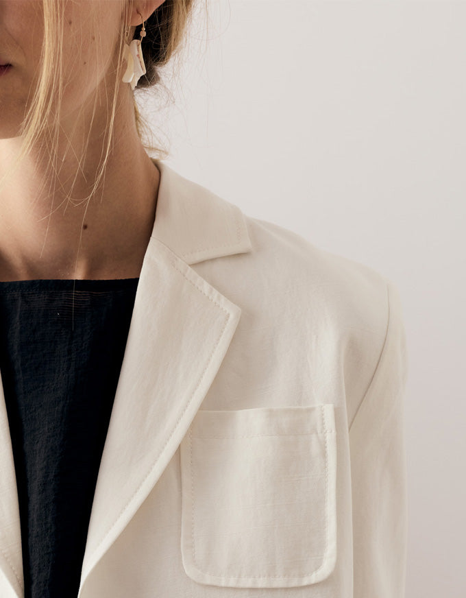 Close-up of a person wearing a light cream or off-white blazer over a dark navy top, with a plain light background.