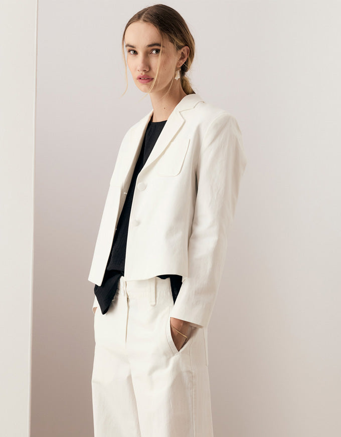 A woman modeling a white tailored blazer and matching trousers over a black top, standing against a plain light beige studio wall.