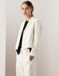 A woman modeling a white tailored blazer and matching trousers over a black top, standing against a plain light beige studio wall.