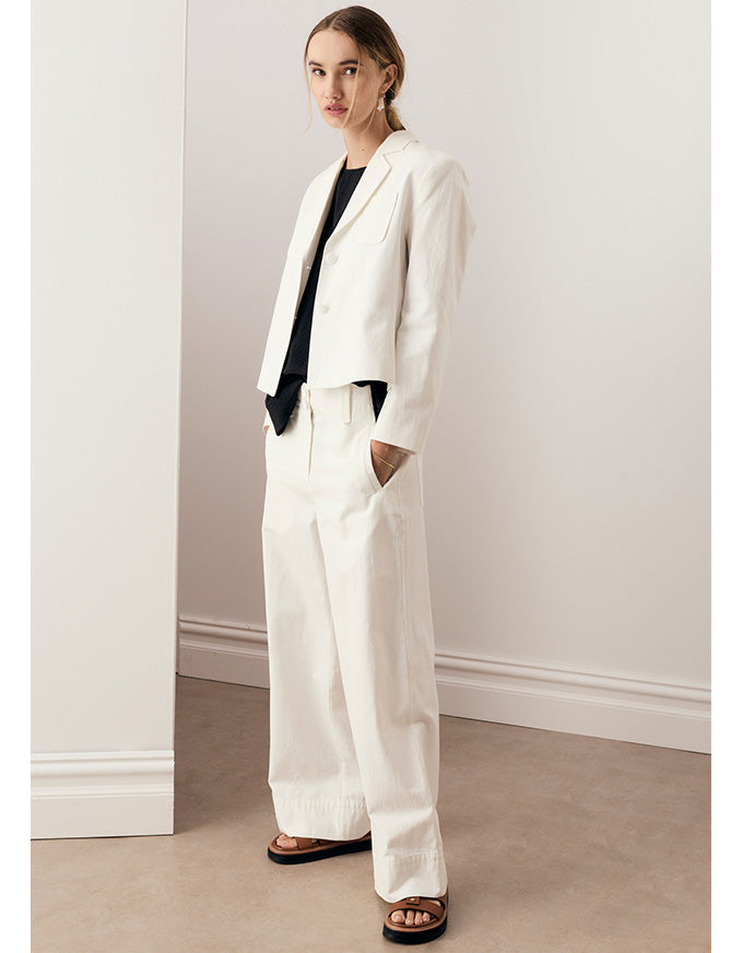A woman models a white tailored blazer and matching wide-leg trousers, styled with a black top and brown slide sandals, standing in a minimalist room with white walls and a beige floor.