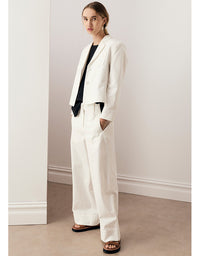 A woman models a white tailored blazer and matching wide-leg trousers, styled with a black top and brown slide sandals, standing in a minimalist room with white walls and a beige floor.