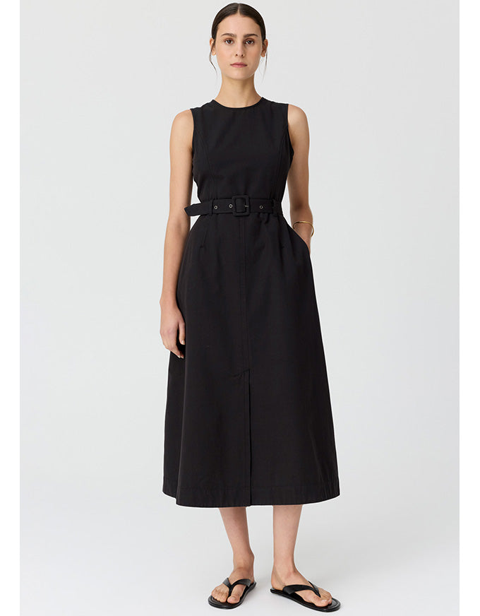 A woman modeling a sleeveless black midi dress with a matching belt, styled with black thong sandals against a light gray studio background.