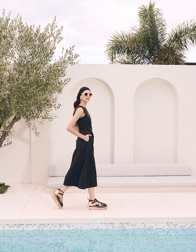 A woman wearing a sleeveless black midi dress and black strappy platform sandals walks beside a swimming pool in front of a white stucco wall with arched recesses and palm trees.