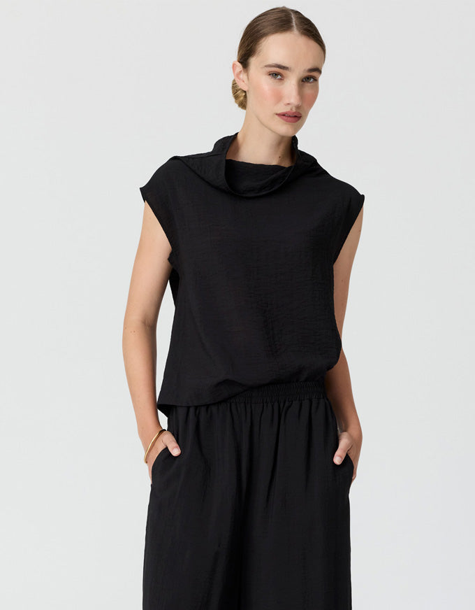 A woman modeling a black sleeveless cowl-neck top with matching black wide-leg trousers, photographed against a light gray studio background.