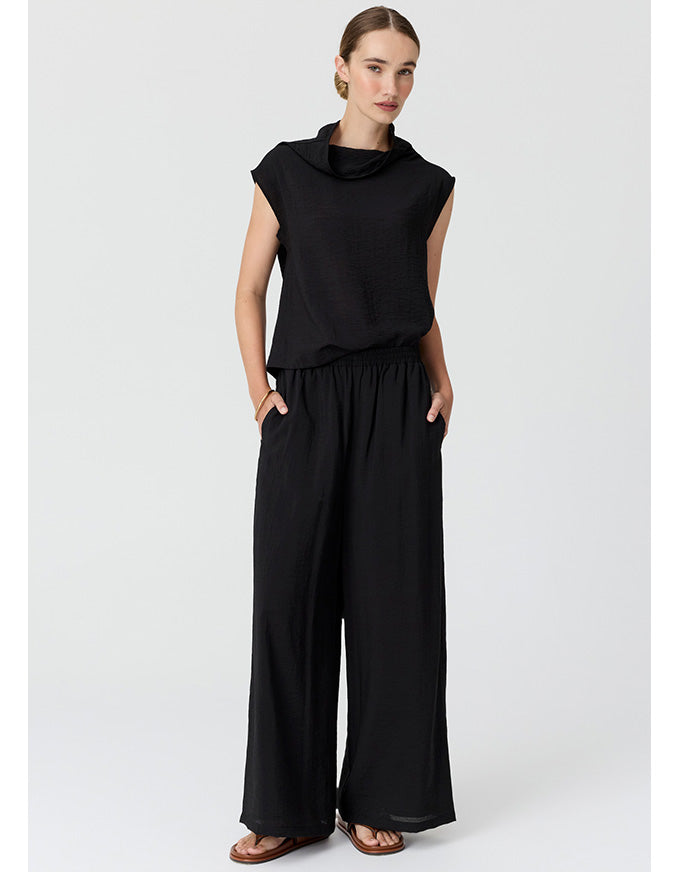 A woman modeling a black sleeveless cowl-neck top and matching black wide-leg pants against a light gray studio background.