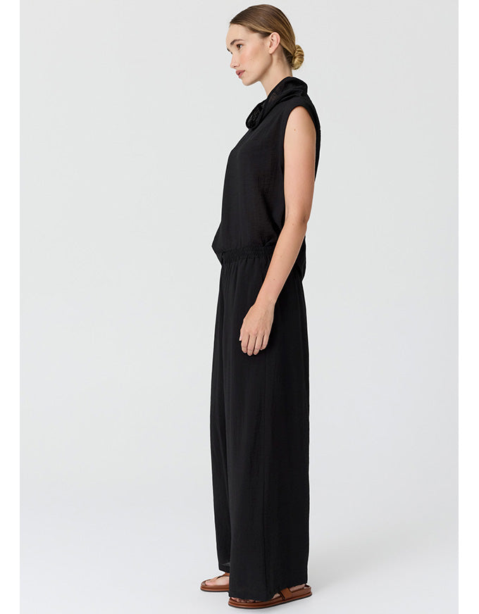 A woman modeling a black sleeveless cowl-neck jumpsuit with wide legs, shown in profile against a light gray studio background.
