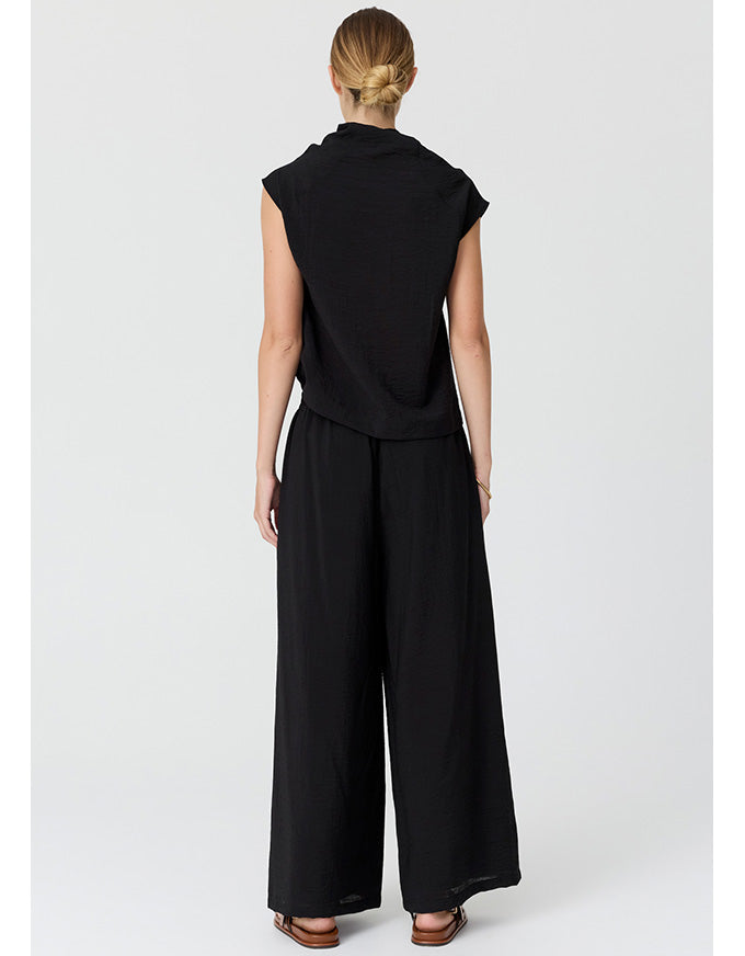 A woman viewed from the back wearing a black sleeveless top and wide-leg black pants, standing against a light gray studio background.