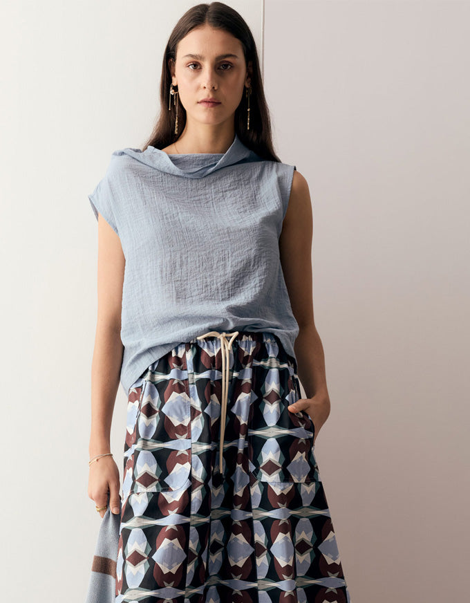 A woman modeling a light blue sleeveless draped top paired with high-waisted patterned pants featuring a brown, white, and gray geometric design with a cream drawstring, set against a plain off-white background.