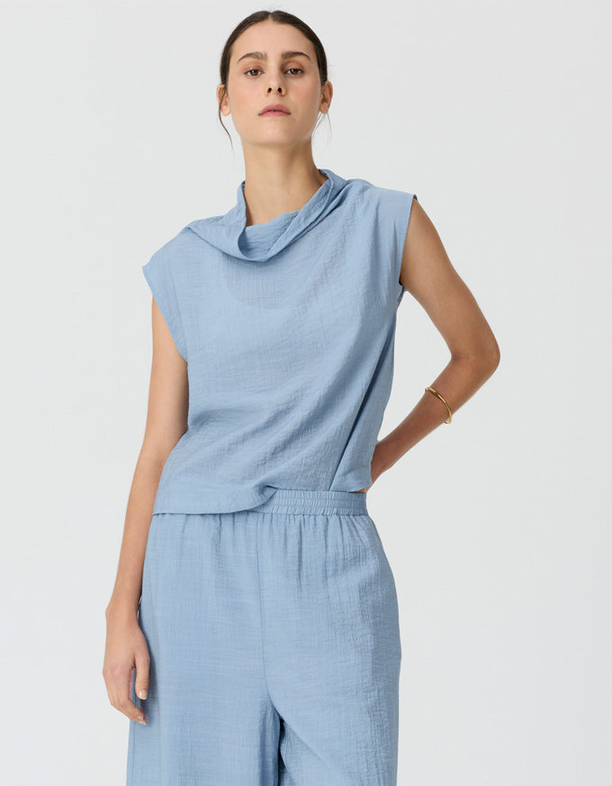 A woman modeling a light blue sleeveless cowl-neck top and matching wide-leg trousers against a plain light gray studio background.