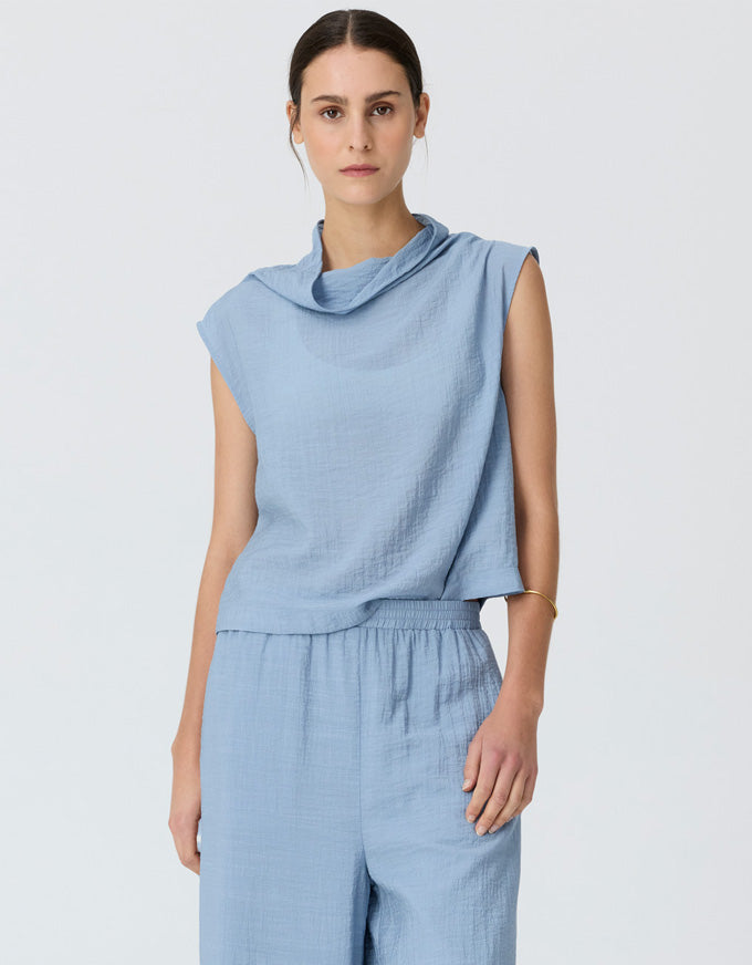 A woman modeling a light blue sleeveless top with a draped cowl neckline and matching light blue wide-leg trousers, photographed against a plain light gray studio background.