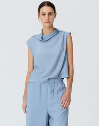 A woman modeling a light blue sleeveless top with a draped cowl neckline and matching light blue wide-leg trousers, photographed against a plain light gray studio background.