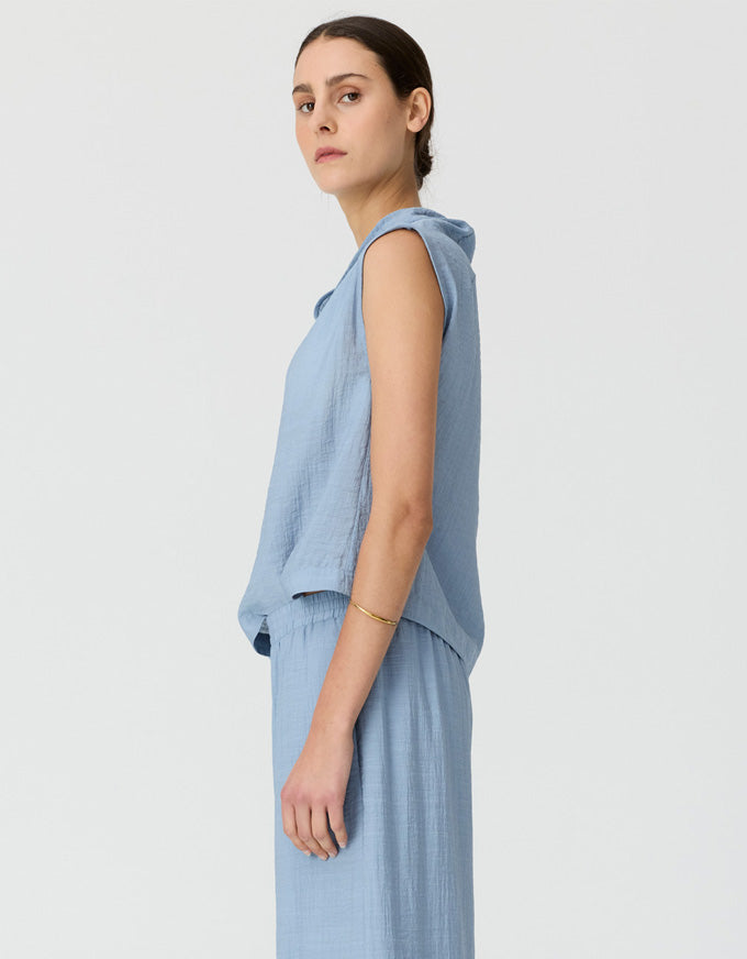 A woman in profile wearing a light blue sleeveless drape top and matching wide-leg trousers against a plain light gray studio background.