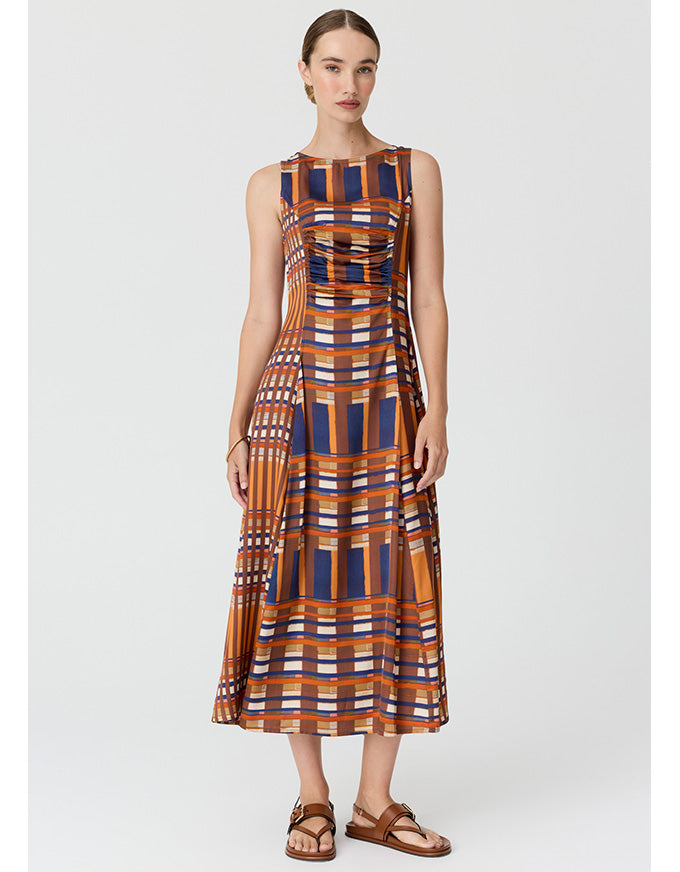 A woman modeling a sleeveless midi dress with a geometric plaid pattern in orange, navy blue, brown, and white, paired with brown leather sandals, against a light gray studio background.