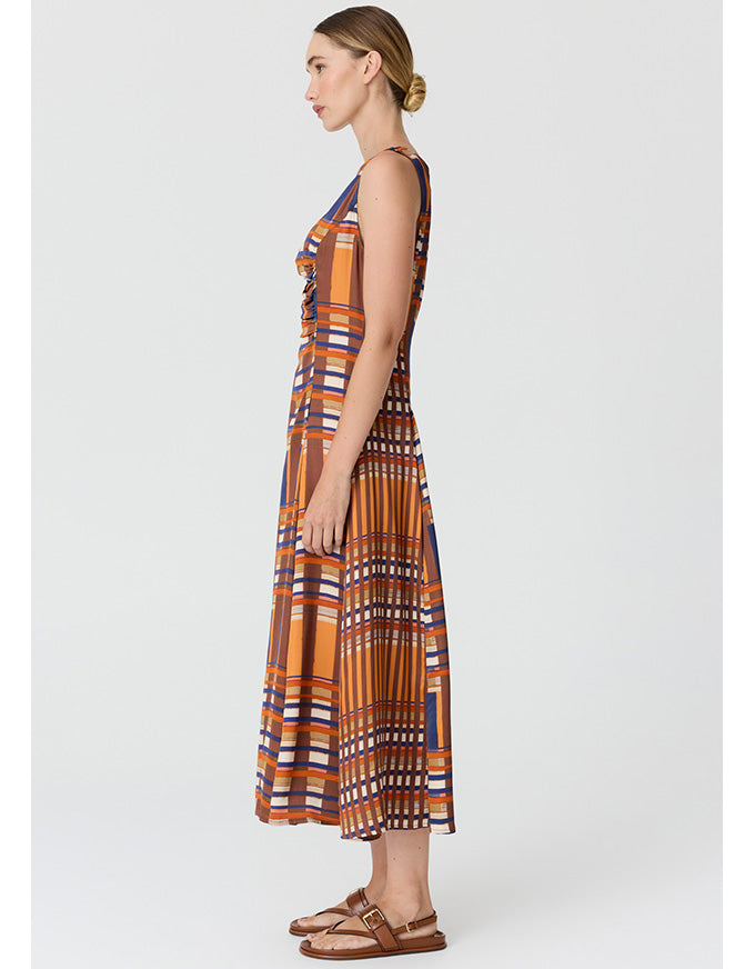 A woman modeling a sleeveless midi dress with a geometric pattern in orange, brown, blue, and white, paired with brown sandals, shown from a side profile against a light gray background.