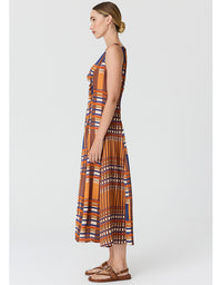 A woman modeling a sleeveless midi dress with a geometric pattern in orange, brown, blue, and white, paired with brown sandals, shown from a side profile against a light gray background.