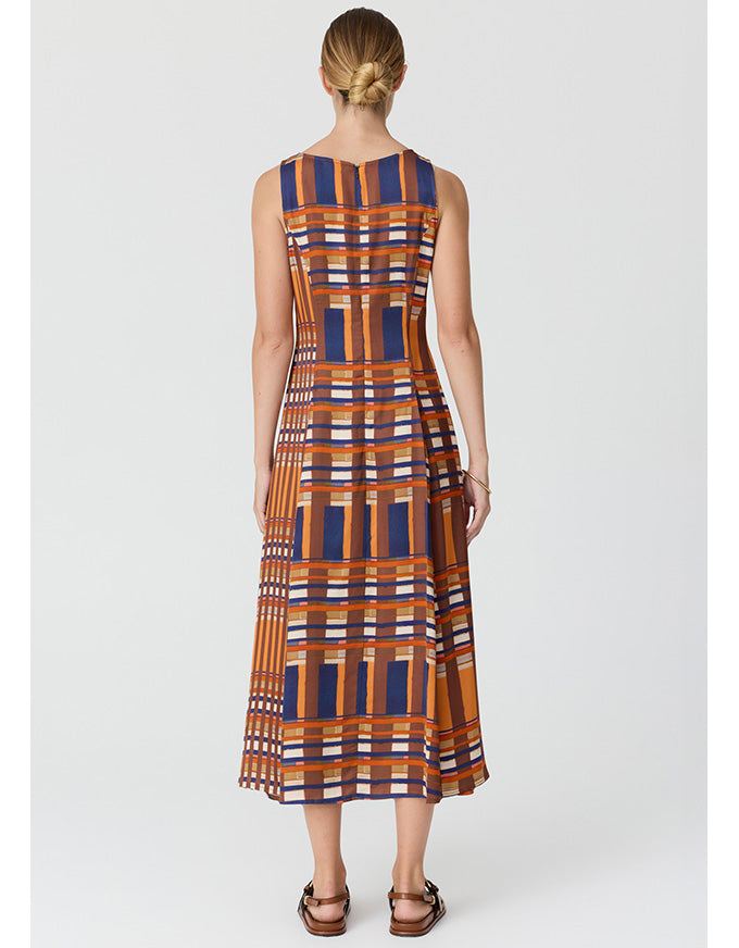 A woman viewed from the back wearing a sleeveless midi dress with a geometric plaid pattern in orange, navy blue, and white, paired with brown leather sandals, against a light gray studio background.