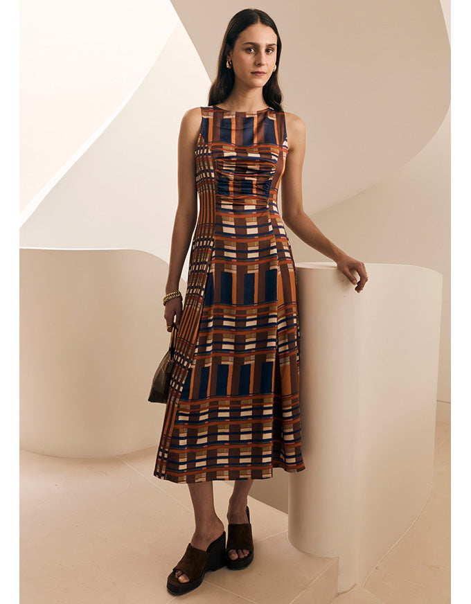 A woman models a sleeveless midi dress with a geometric plaid pattern in brown, navy blue, and beige, standing in a minimalist cream-colored architectural setting.