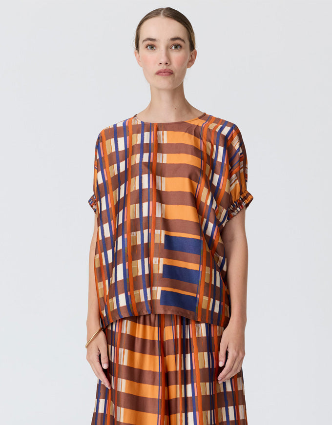 A model wearing a loose-fitting short-sleeve top and matching pleated skirt set with a bold orange, brown, navy blue, and white striped pattern, shot against a light gray studio background.