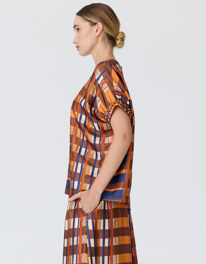 A woman in profile wearing a matching two-piece plaid set with short puff sleeves and a high neckline, featuring orange, brown, blue, and white stripes, against a light gray studio background.