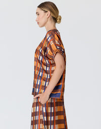 A woman in profile wearing a matching two-piece plaid set with short puff sleeves and a high neckline, featuring orange, brown, blue, and white stripes, against a light gray studio background.