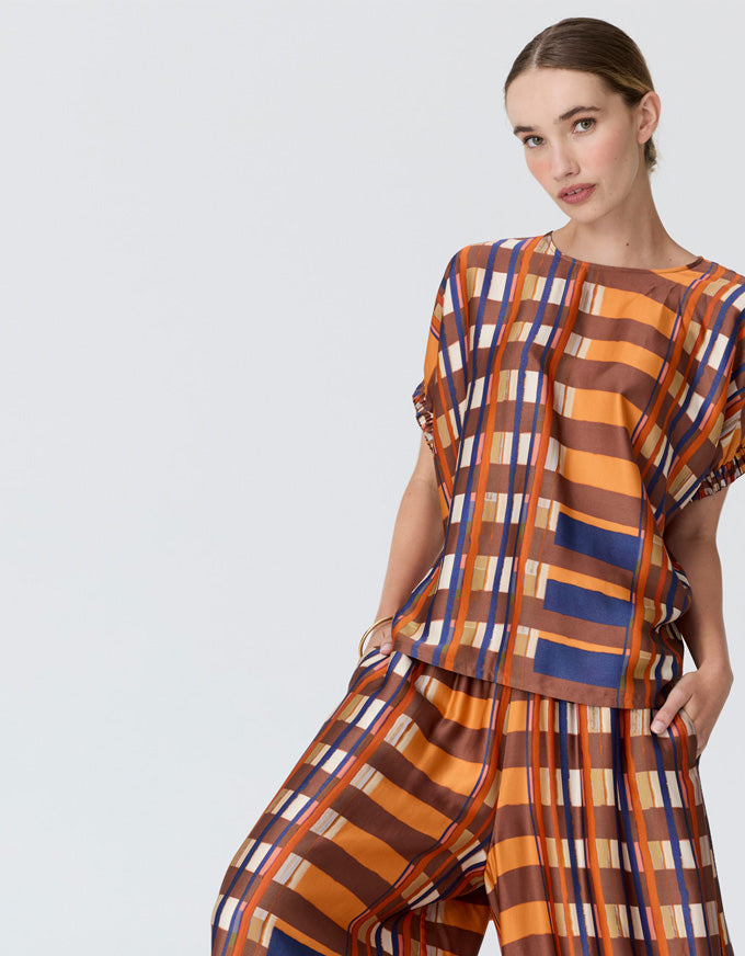 A woman modeling a matching two-piece outfit with a bold vertical stripe pattern in orange, brown, blue, and white, photographed against a light gray studio background.